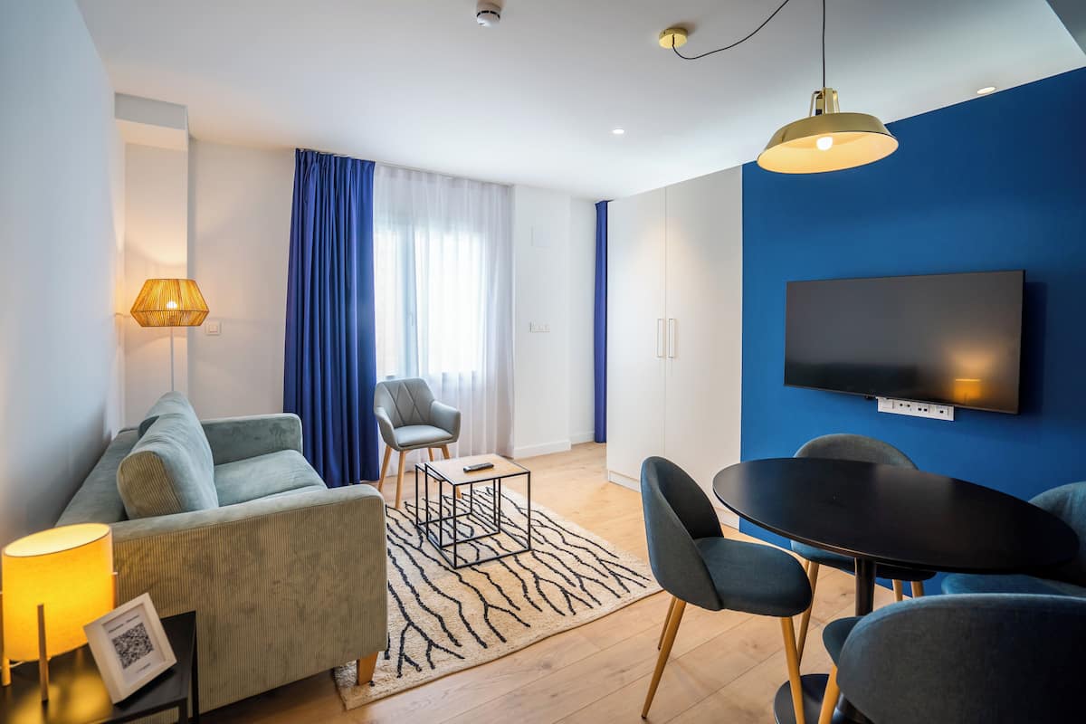 Een moderne woonkamer met een bank, een eettafel, stoelen, een aan de muur bevestigde televisie en een groot raam met gordijnen.