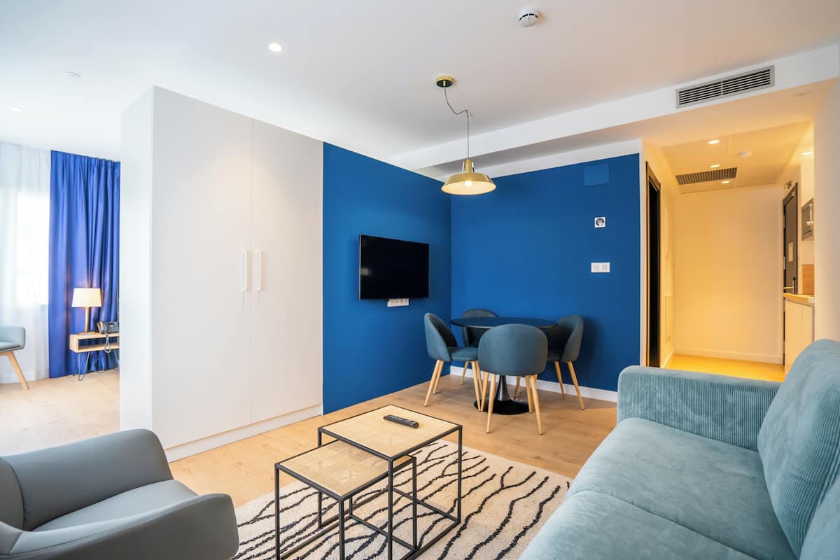 Een moderne woonkamer met een blauwe accentmuur, een flatscreen televisie, een eethoek met een tafel en stoelen, en een bank.