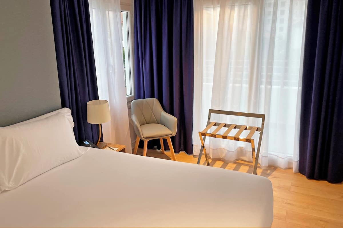 Een hotelkamer met een groot bed, een stoel, een klein tafeltje en een raam met gordijnen.