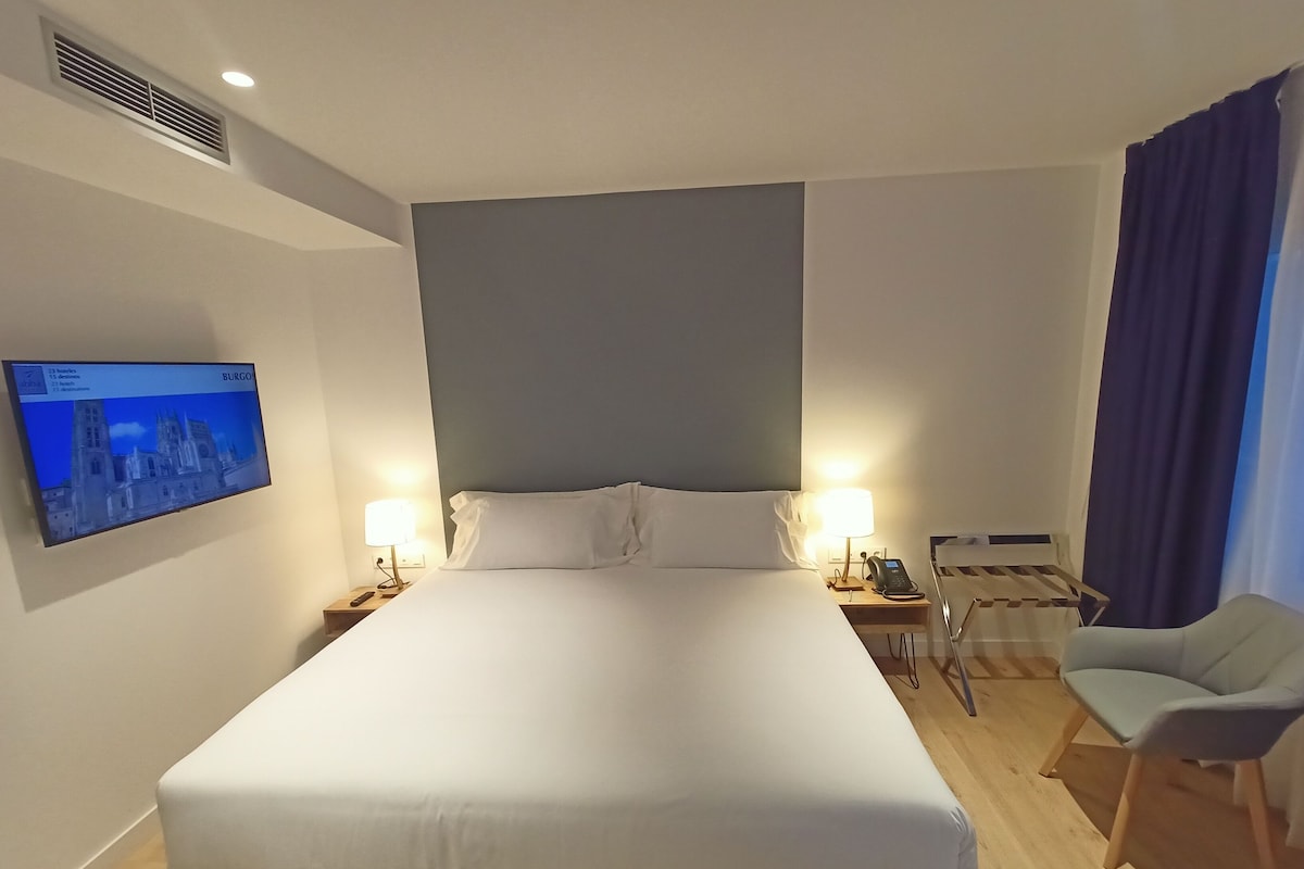 Ein Hotelzimmer mit einem großen Bett, zwei Nachttischen mit Lampen, einem Flachbildfernseher an der Wand und einem Sessel.