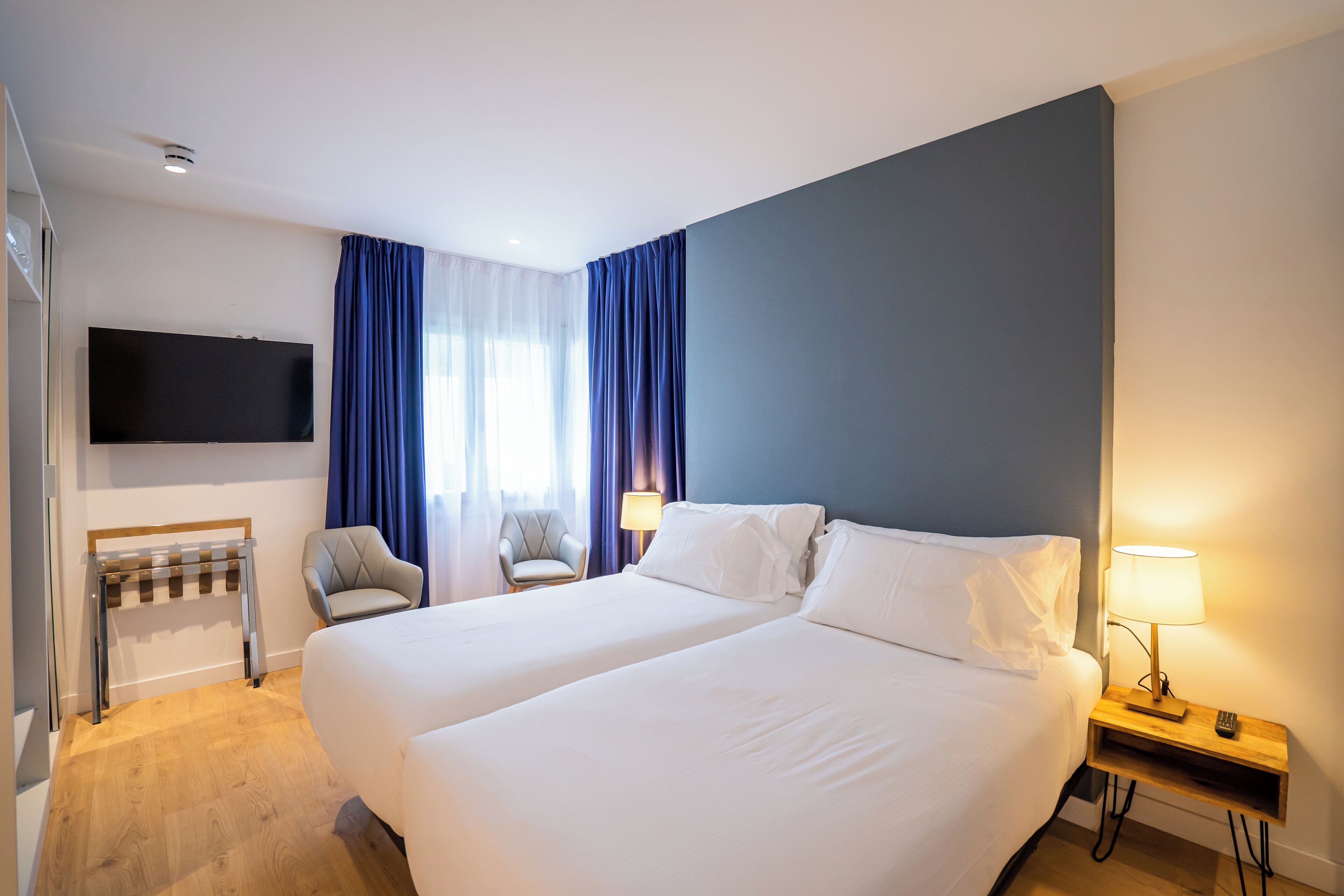 Une chambre d’hôtel avec deux lits, un téléviseur fixé au mur, une chaise, une lampe et une fenêtre avec des rideaux.