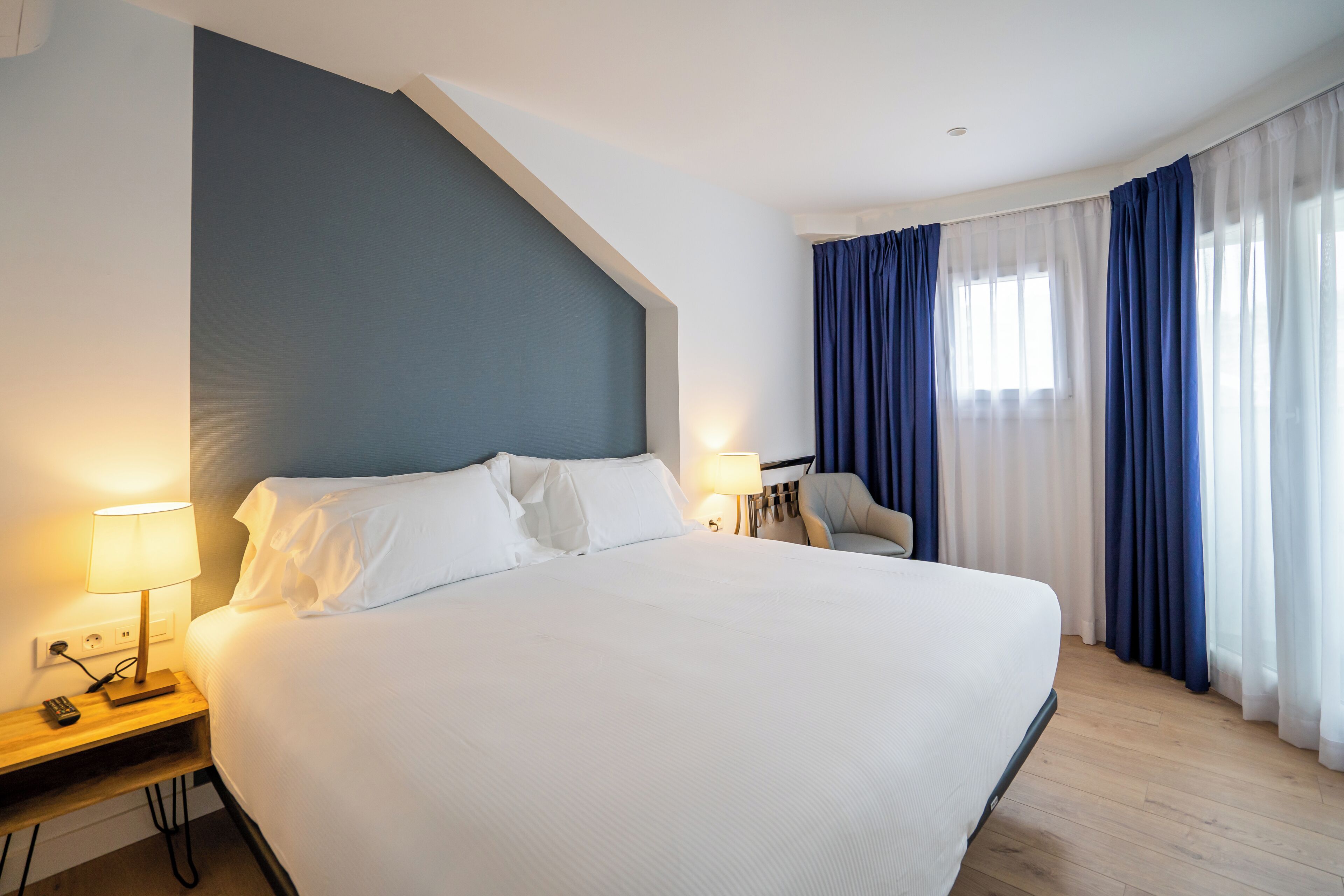 Habitación de hotel con una cama grande, una mesita de noche con lámpara, una silla y un ventanal con cortinas azules.