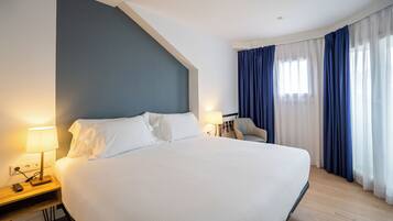 Habitación de hotel con una cama grande, una mesita de noche con lámpara, una silla y un ventanal con cortinas azules.