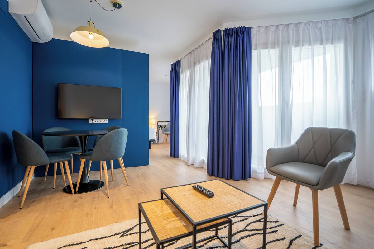 Een moderne woonkamer met een flatscreen televisie, een eettafel met stoelen, een bank en een groot raam met gordijnen.