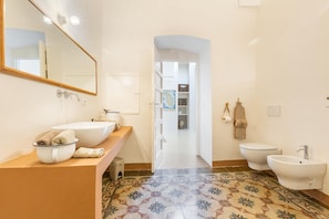 A bathroom with a large mirror, a sink, a bidet, and a toilet. There is a patterned tile floor and a hallway leading to a room with shelves. - 3113 Volte Sotto le Stelle by Barbarhouse (Casarano)