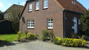 A two-story brick house with a tiled roof, a front door, and a small garden with yellow flowers. - Das Ferienhaus "lennox" mit 70 qm Wohnfläche Bietet Ausreichend Platz für bis zu (Carolinensiel)