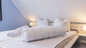 A neatly made bed with white bedding and pillows, a bedside table with a lamp, and a wall-mounted light fixture. - Das Ferienhaus "lennox" mit 70 qm Wohnfläche Bietet Ausreichend Platz für bis zu (Carolinensiel)