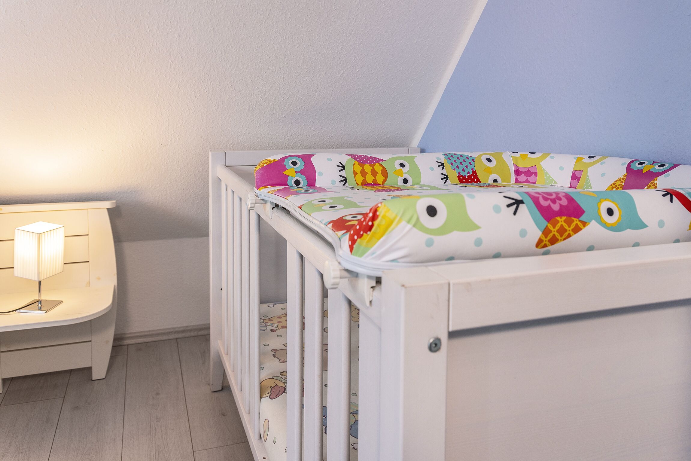 En vit spjälsäng med färgglatt sänglinne med ugglemotiv, ett sängbord med en lampa och ett ljust golv.