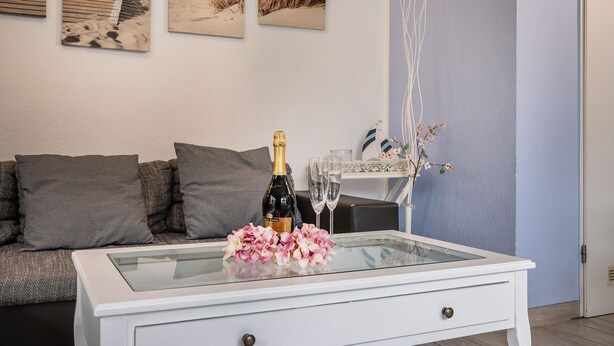 A modern living room with a glass-top coffee table, a bottle of champagne, and a vase of flowers. - Das Ferienhaus "lennox" mit 70 qm Wohnfläche Bietet Ausreichend Platz für bis zu (Carolinensiel)