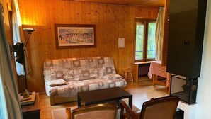 Una habitación acogedora con paredes de madera, un sofá estampado, una mesita, dos sillas, un televisor y una ventana con cortinas.