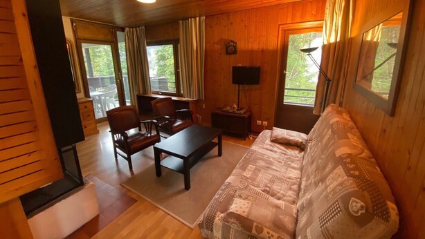 Interior acogedor de una cabaña con techo de madera, un sofá, una mesa de comedor con sillas, un televisor y ventanas grandes que ofrecen vistas al exterior.