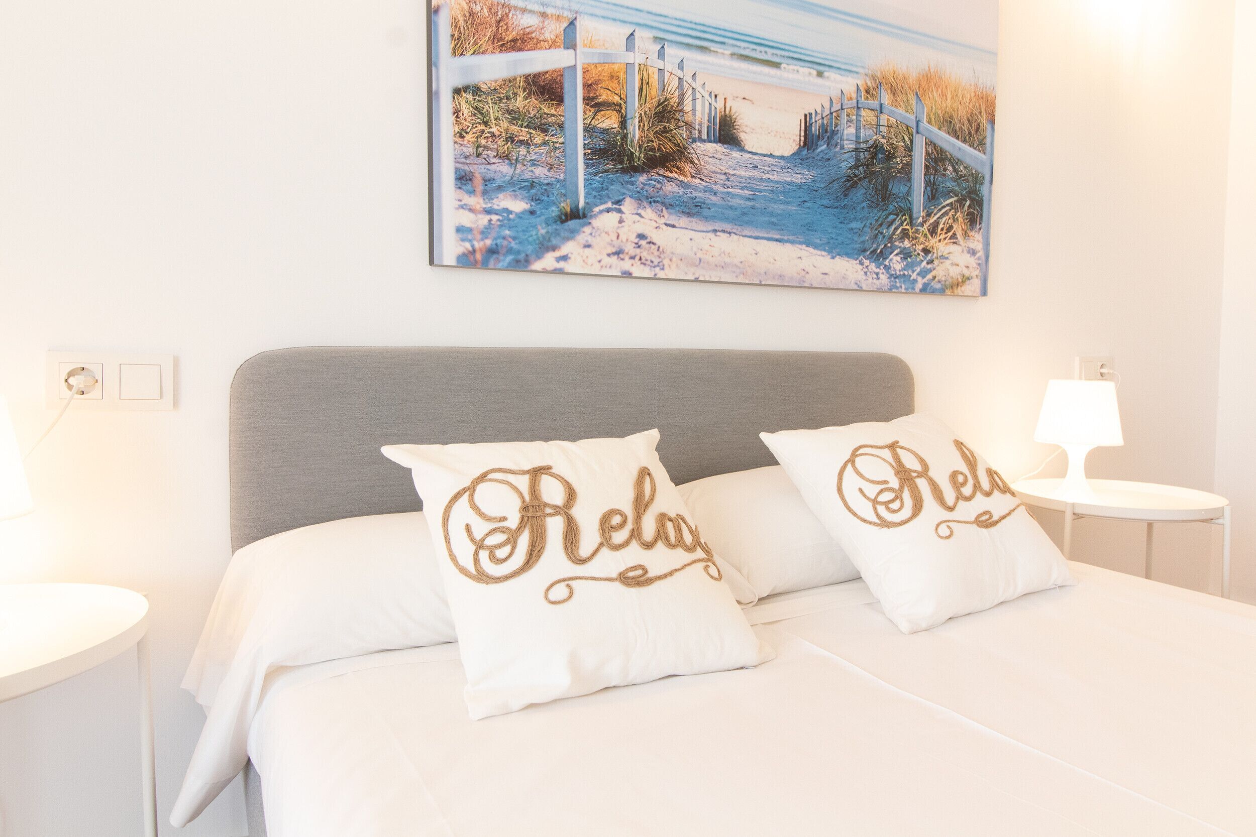 Ein ordentlich bezogenes Bett mit zwei Kissen, ein Gemälde mit Strandmotiv und eine Nachttischlampe.