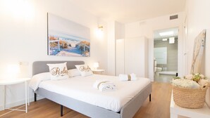 A modern bedroom with a large bed, white bedding, a painting above the bed, and a bathroom visible through an open door.