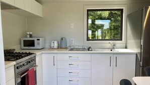 Kompakti keittiö, jossa on valkoiset kaapit, liesi, mikroaaltouuni, vedenkeitin ja ikkuna, josta avautuu näkymä vehreään maisemaan.