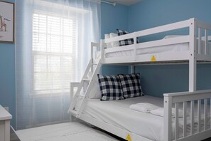 Una cama litera con colchón y almohadas, una ventana con cortinas traslúcidas y un cuadro colgado en la pared.