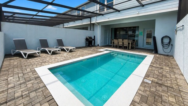 A rectangular swimming pool with a tiled deck, surrounded by lounge chairs and a covered patio area. - Beautiful 5-Bedroom Home with Pool (Four Corners)
