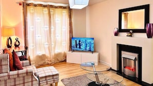 A living room with a sofa, ottoman, TV, glass table, fireplace, and curtains. - Lovely 2-bed Apartment in Aberdeen (Aberdeen)
