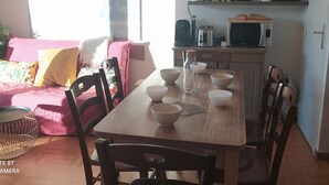 Ett mysigt vardagsrum med ett matbord i trä för fyra personer, en rosa soffa med blommiga kuddar och en köksdel med vattenkokare, brödrost och mikrovågsugn.