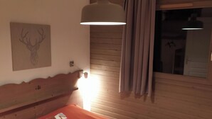 Ett sovrum med en sänggavel i trä, en säng med ett orange överkast, en vägglampa, ett fönster med gardiner och en väggdekoration som föreställer ett rådjurshuvud.