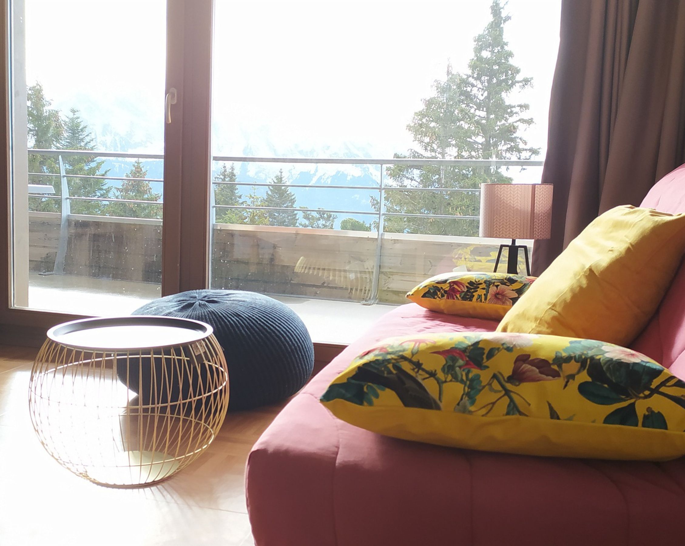 A room with a large window, a pink sofa with floral cushions, a round wooden chair, and a small table.