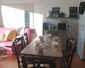 Un salón acogedor con una mesa de comedor de madera para cuatro, un sofá rosa con cojines estampados y una zona de cocina con hervidor, tostadora y microondas.