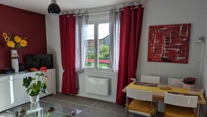 A modern living room with a glass coffee table, white furniture, a red and white curtain, a red abstract painting, and a view of greenery outside. - Charming apartment with terrace and garden, perfect for 4 people, in Barbezieux (Barbezieux-Saint-Hilaire)