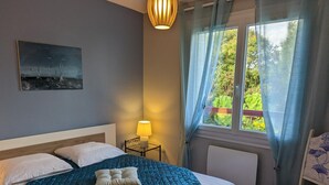A bedroom with a bed, a bedside lamp, a chair, a window with curtains, and a painting on the wall. - Charming apartment with terrace and garden, perfect for 4 people, in Barbezieux (Barbezieux-Saint-Hilaire)