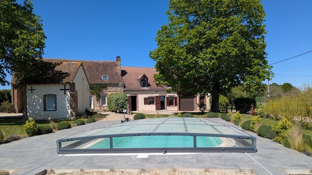 A swimming pool with a glass enclosure, surrounded by a pebble beach, with houses and trees in the background. - Cozy Countryside Cottage with Spacious Garden – Sleeps 4-5 (Douchy-Montcorbon-Douchy)