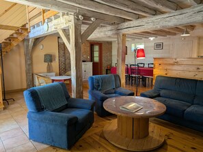 Un intérieur rustique avec des poutres apparentes, une table ronde en bois, des canapés bleus et un coin repas avec une nappe rouge.