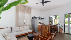 A modern living room with a sofa, wooden chairs, a coffee table, a kitchen area with a refrigerator, and a large window with curtains. - Amazing Villa in la Veleta 14 pax (Tulum)