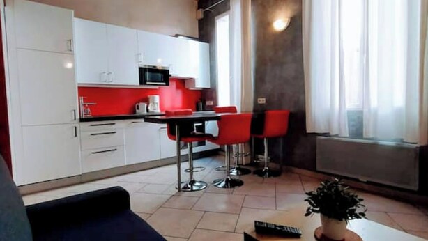 Een moderne keuken met witte kasten, een rode aanrecht en een magnetron. Er zijn twee rode barkrukken en een eettafel. De kamer heeft een groot raam met gordijnen en een plant op het raamkozijn.