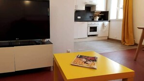 Un saló amb un televisor de pantalla plana sobre un moble, una taula groga amb una revista, armaris blancs, una zona de cuina amb fogó i forn, i una finestra amb cortines.