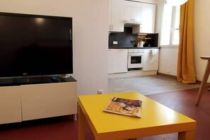 A living room with a flat-screen TV on a stand, a yellow table with a magazine, white cabinets, a kitchen area with a stove and oven, and a window with curtains.