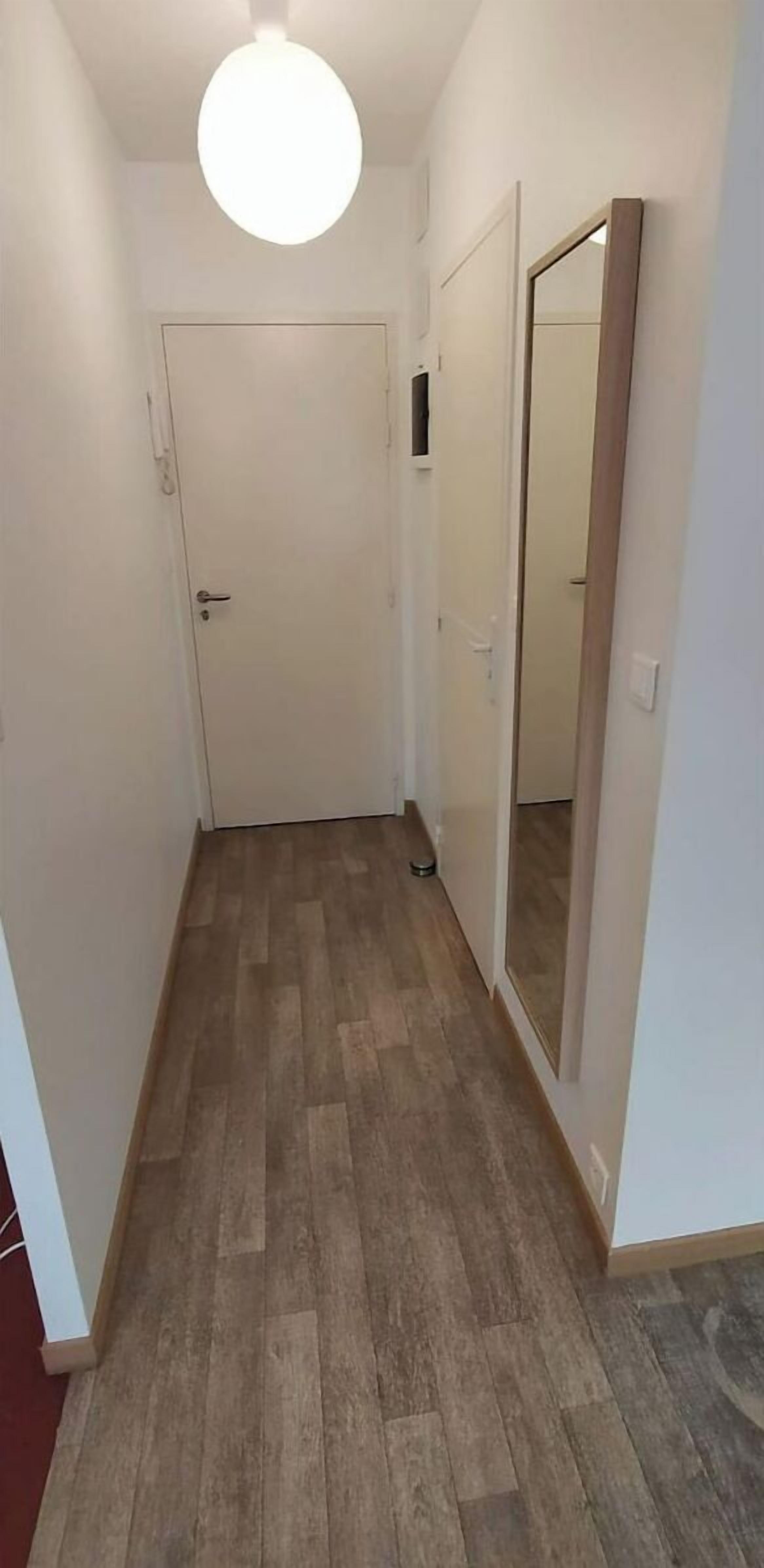 A narrow hallway with a wooden floor, a white door, and a mirror on the right wall.