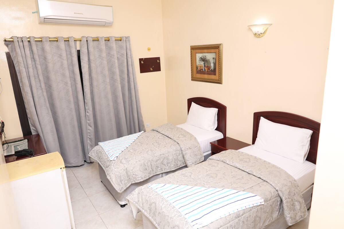 A hotel room with two beds, a desk, an air conditioner, a wall-mounted picture, and a curtain.