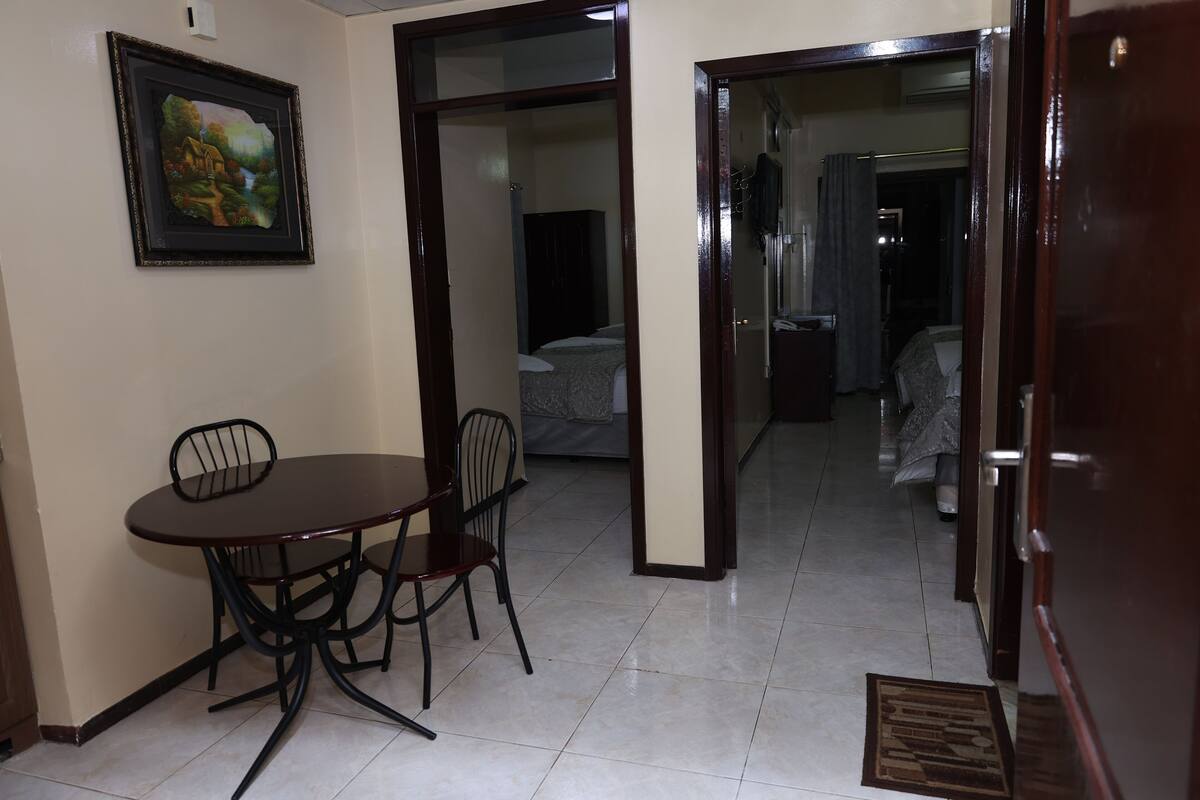 A room with a dining table and chairs, a bed with a gray bedspread, and a wardrobe.