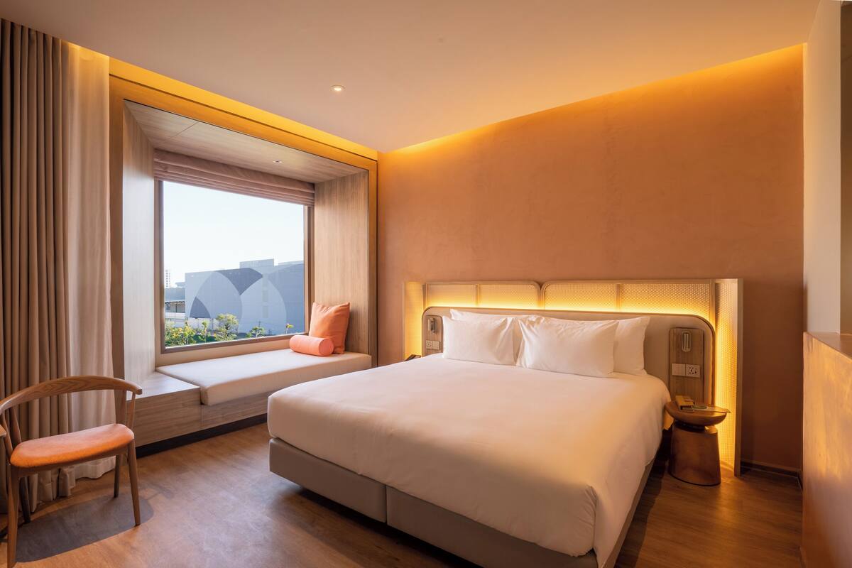 Habitación de hotel con una cama grande, un asiento junto a la ventana, una mesita de noche y vistas a edificios.