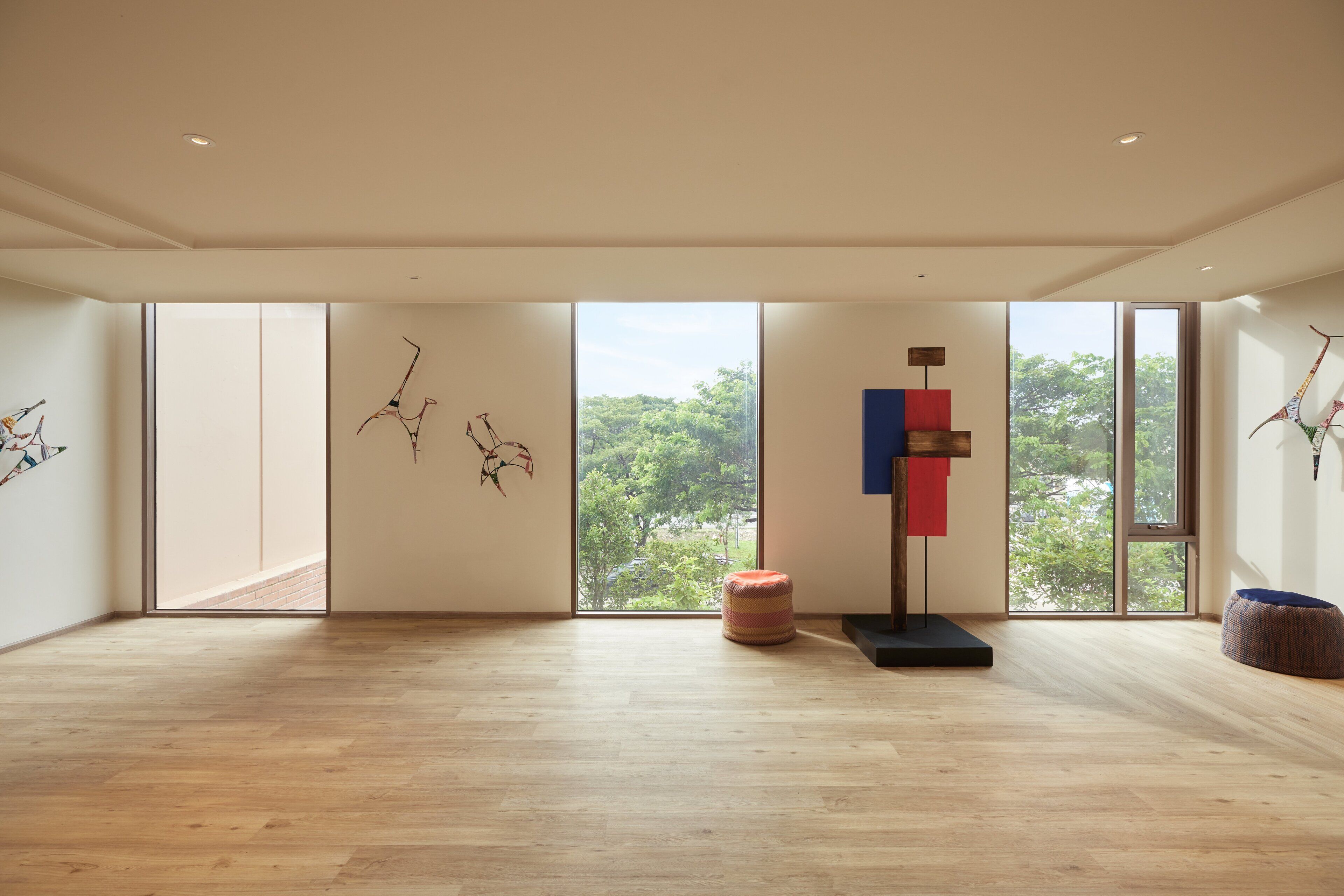 Una habitación amplia con grandes ventanales, piso de madera y decoración minimalista que incluye un cuadro abstracto de pared y algunos otomanes.