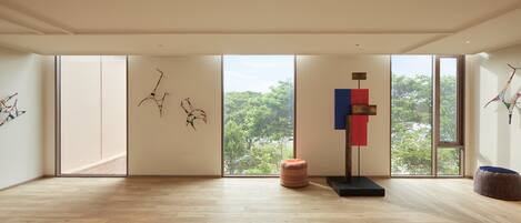 Una habitación amplia con grandes ventanales, piso de madera y decoración minimalista que incluye un cuadro abstracto de pared y algunos otomanes.