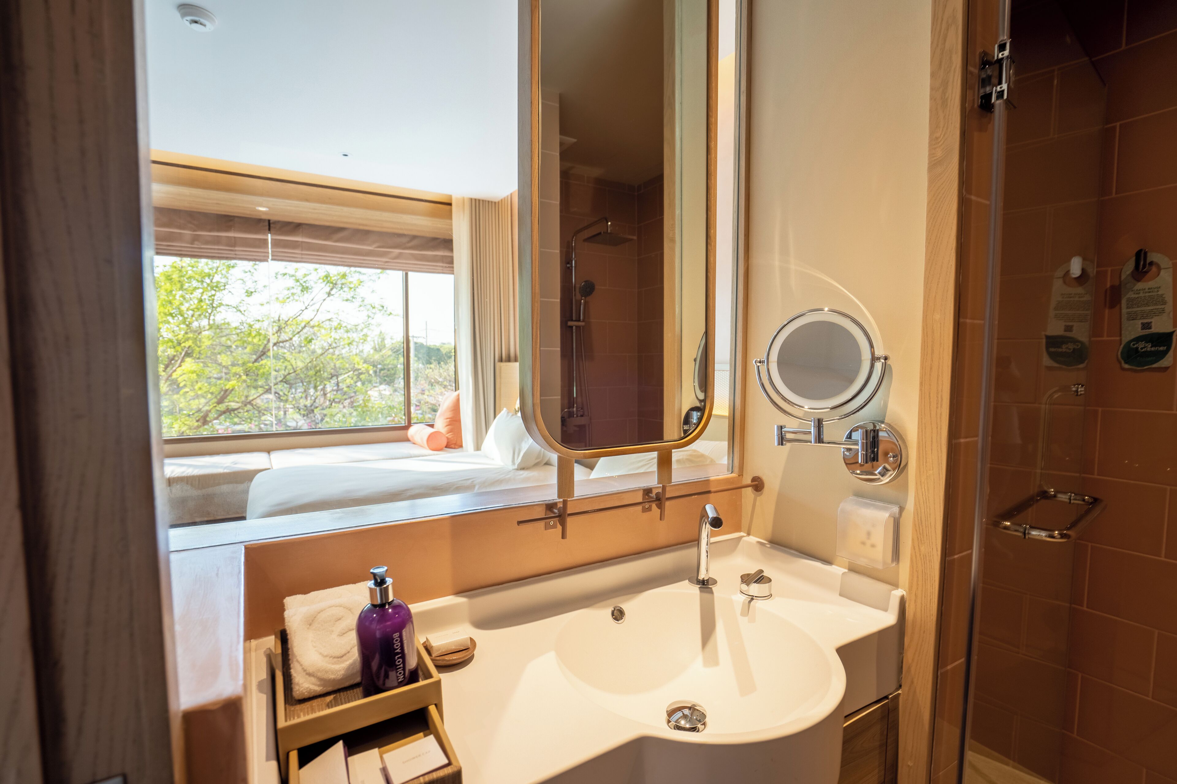 Une salle de bain avec un grand miroir, un lavabo, une douche et une fenêtre donnant sur les arbres.
