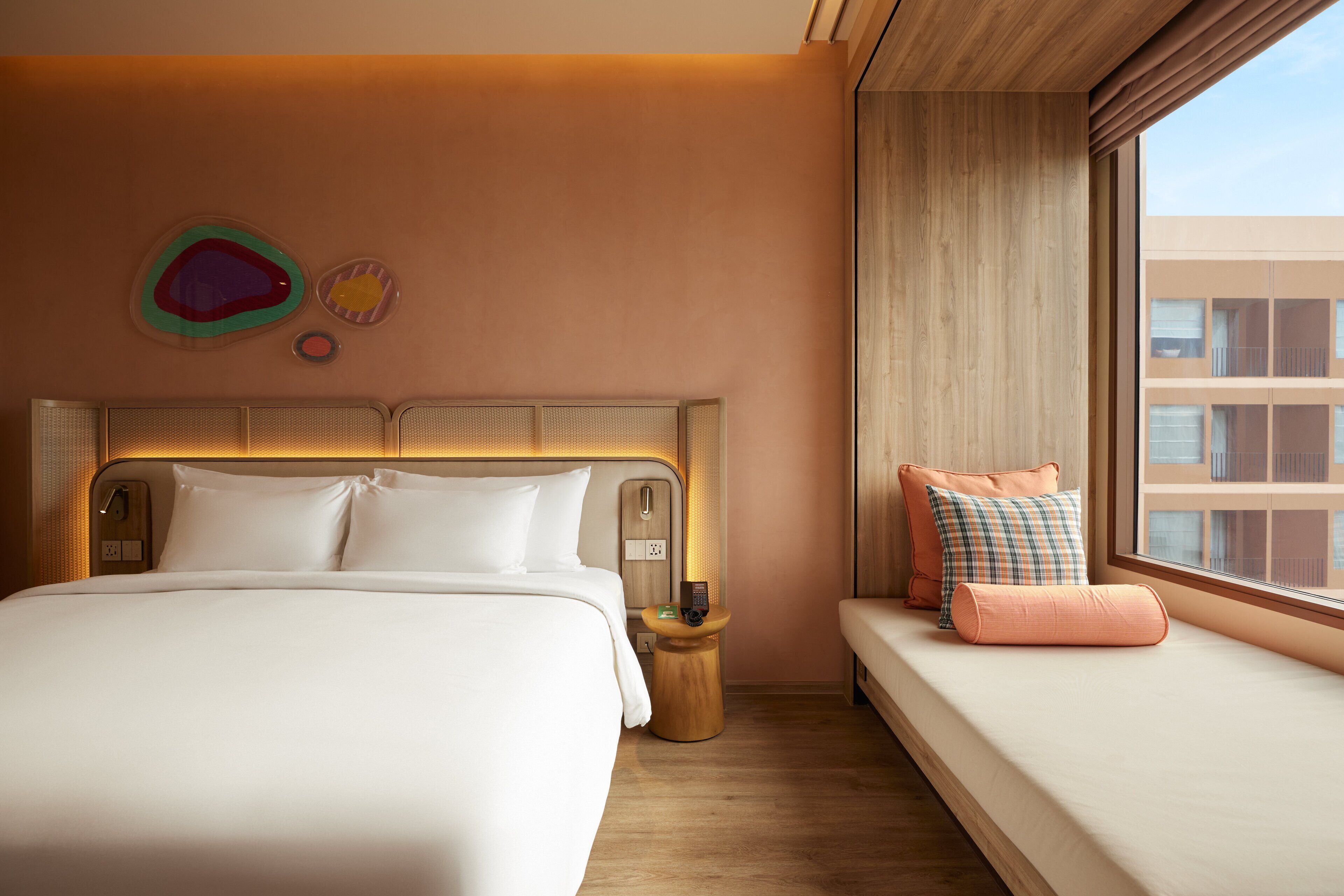 A hotel room with a large bed, a bedside table, a small table with a remote control, and a window with a view of buildings.