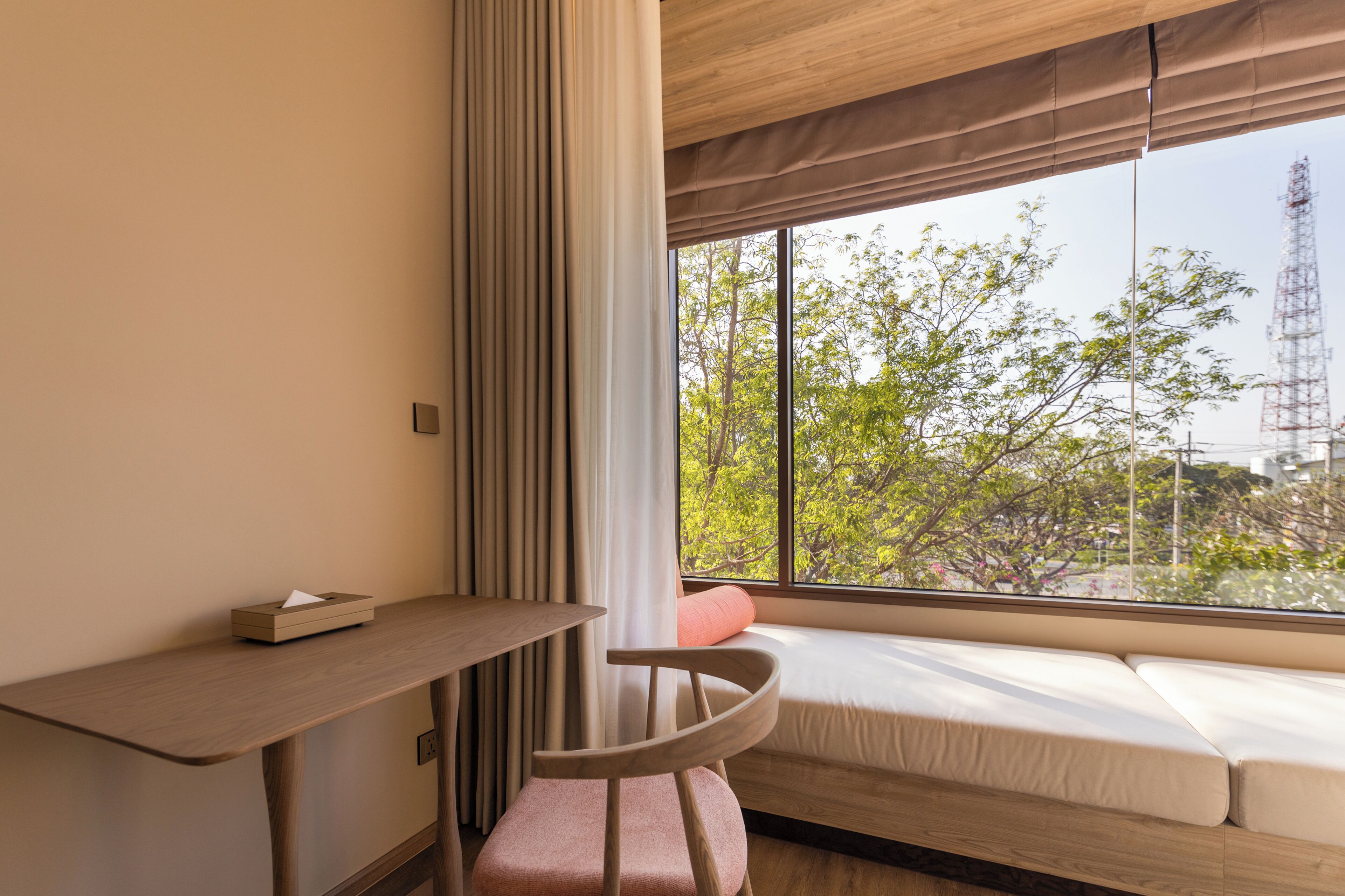 Una habitación con un escritorio de madera, una silla con reposabrazos, un sofá y un ventanal con vistas a árboles y una torre.