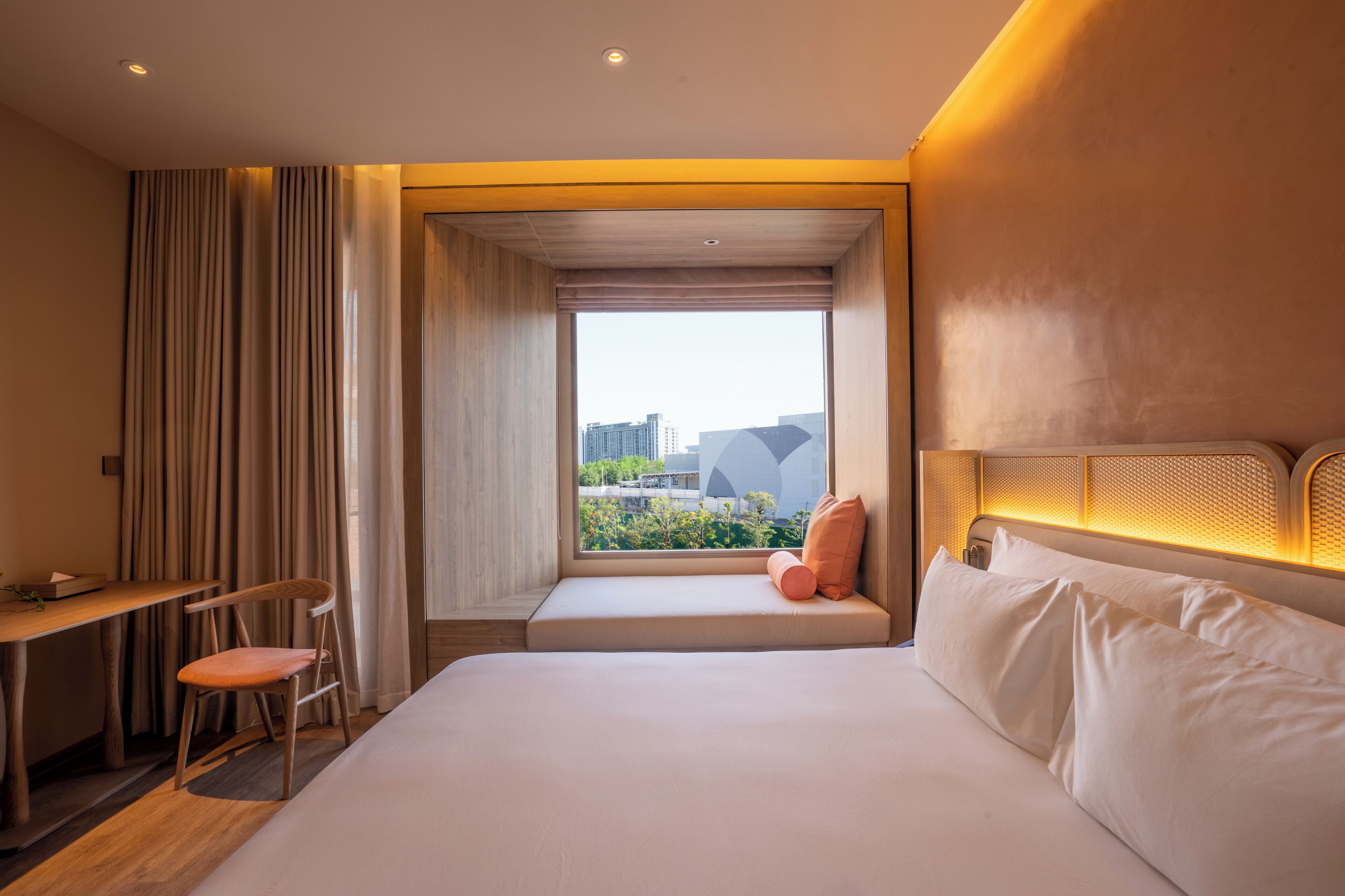 A hotel room with a bed, a desk, a chair, a window with a view of buildings, and a headboard with built-in lighting.