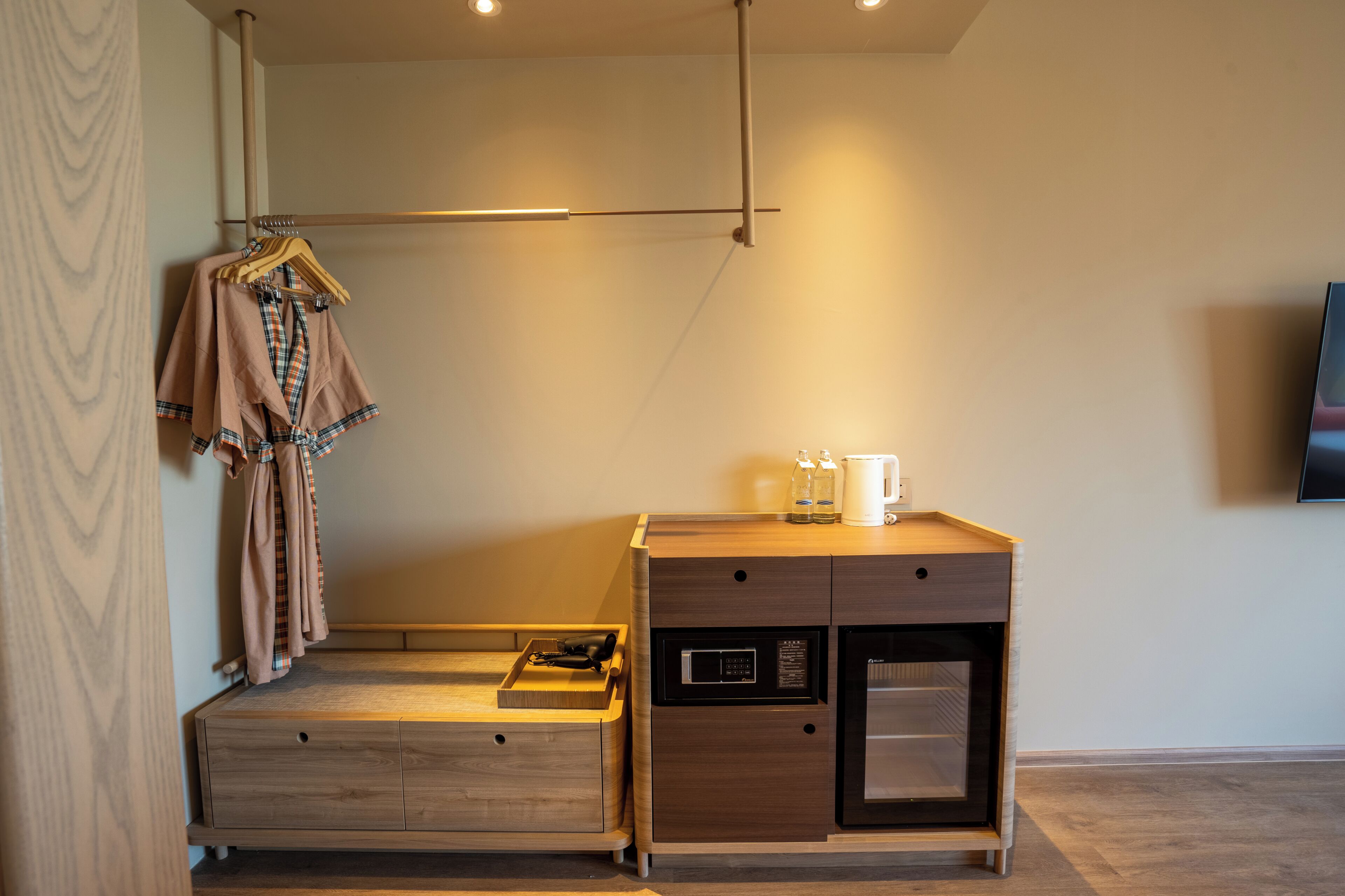 Una habitación compacta con una cama de madera, un escritorio pequeño con hervidor, microondas y televisor montado en la pared.