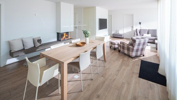 Uma sala de estar moderna com uma mesa de jantar de madeira, cadeiras brancas, uma poltrona xadrez e uma lareira.