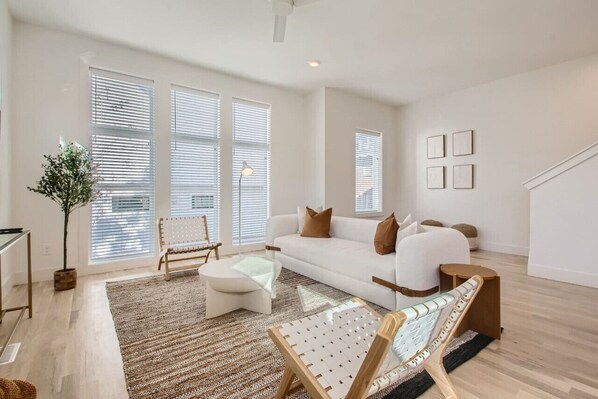 A spacious living room with a white sofa, a coffee table, and a striped rug. There are large windows with blinds, a potted plant, and a bench.