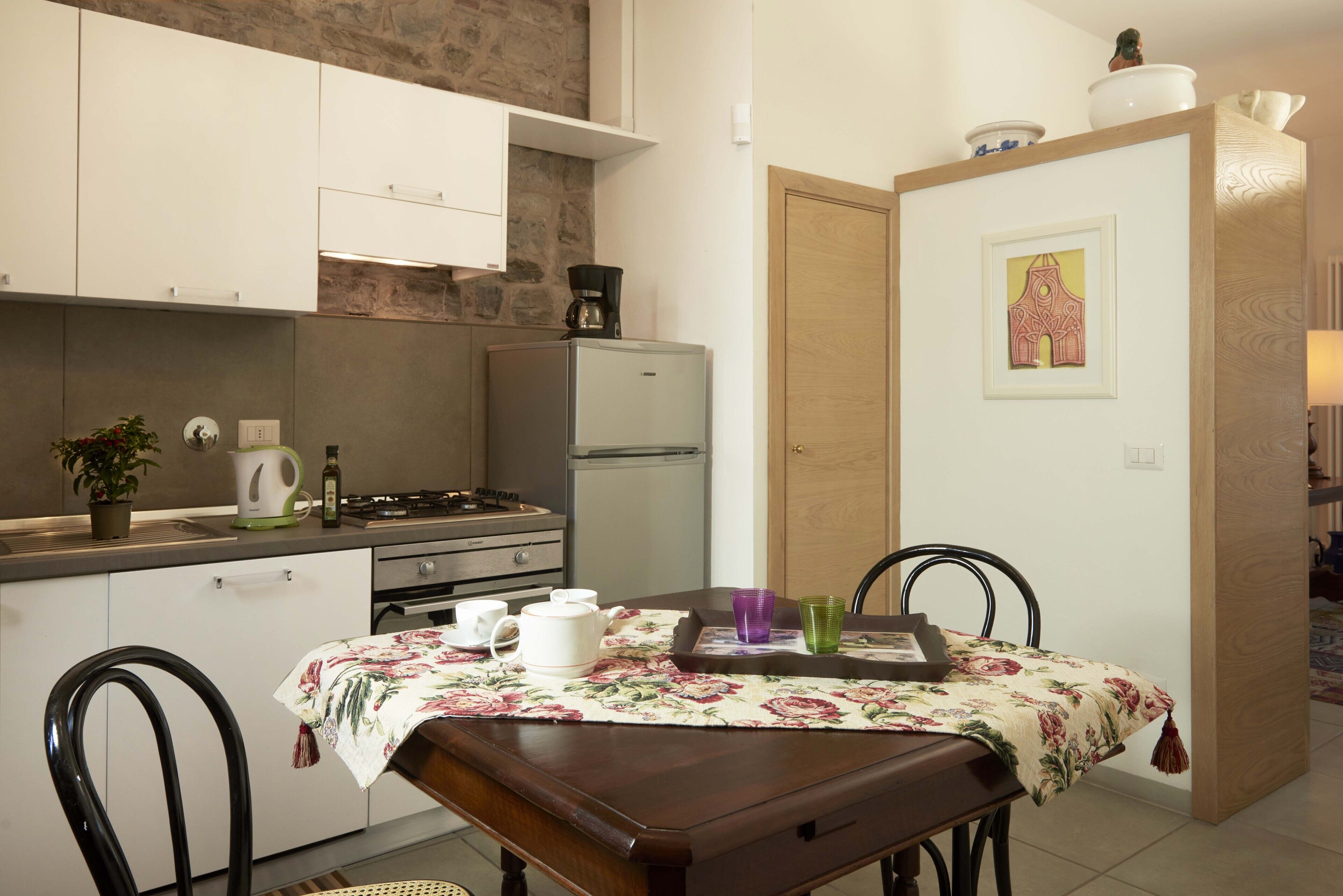 A compact kitchen with white cabinets, a refrigerator, a stove, and a dining table with a floral tablecloth.