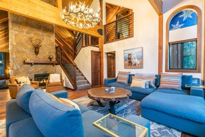 A spacious living room with a stone fireplace, a chandelier, a staircase, and a large window with a view of trees.