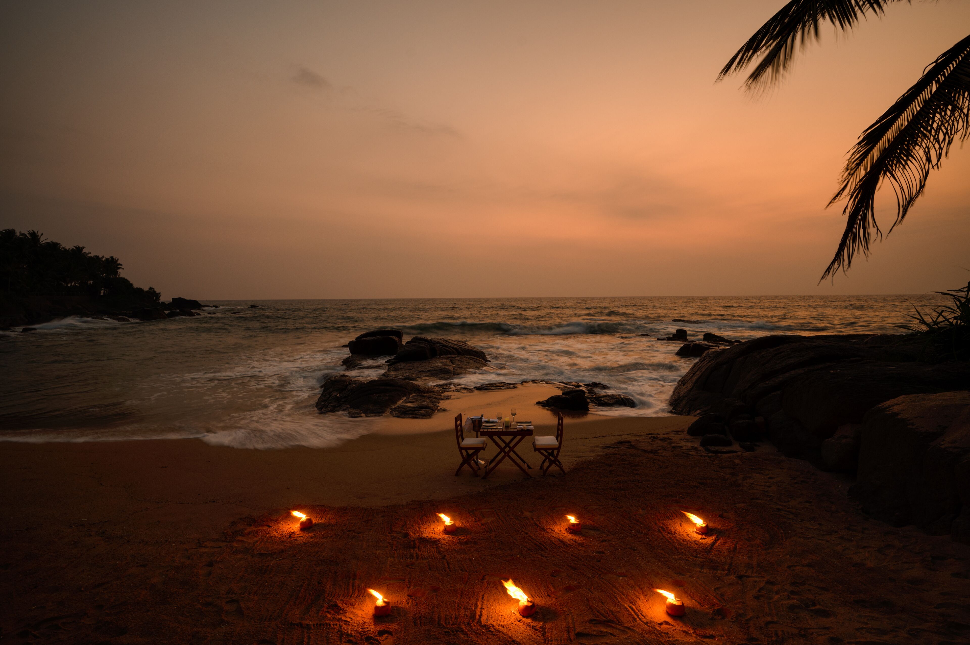 A beach scene with a table and chairs set up on the sand, surrounded by lit candles.