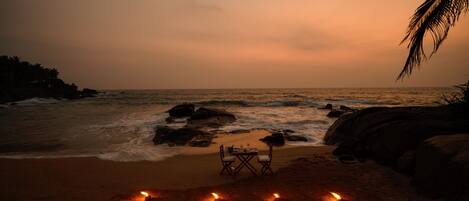 A beach scene with a table and chairs set up on the sand, surrounded by lit candles.